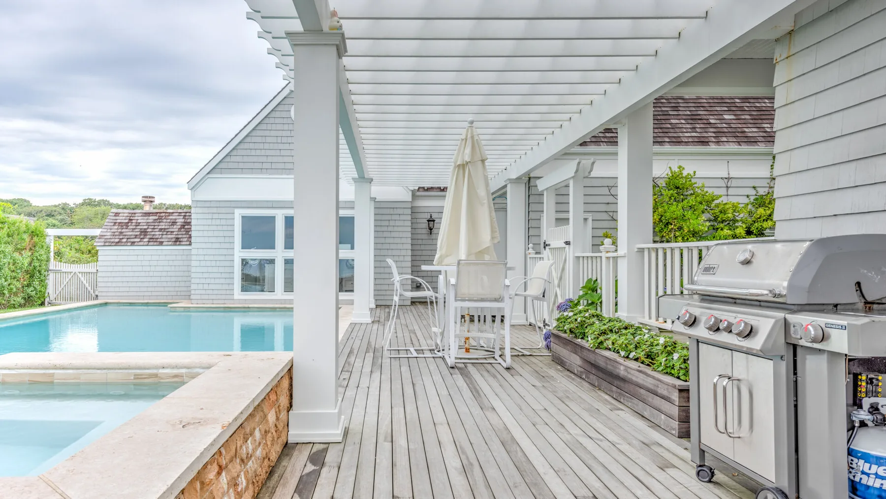 A covered outdoor deck overlooks a swimming pool, with dining seating and a grill.