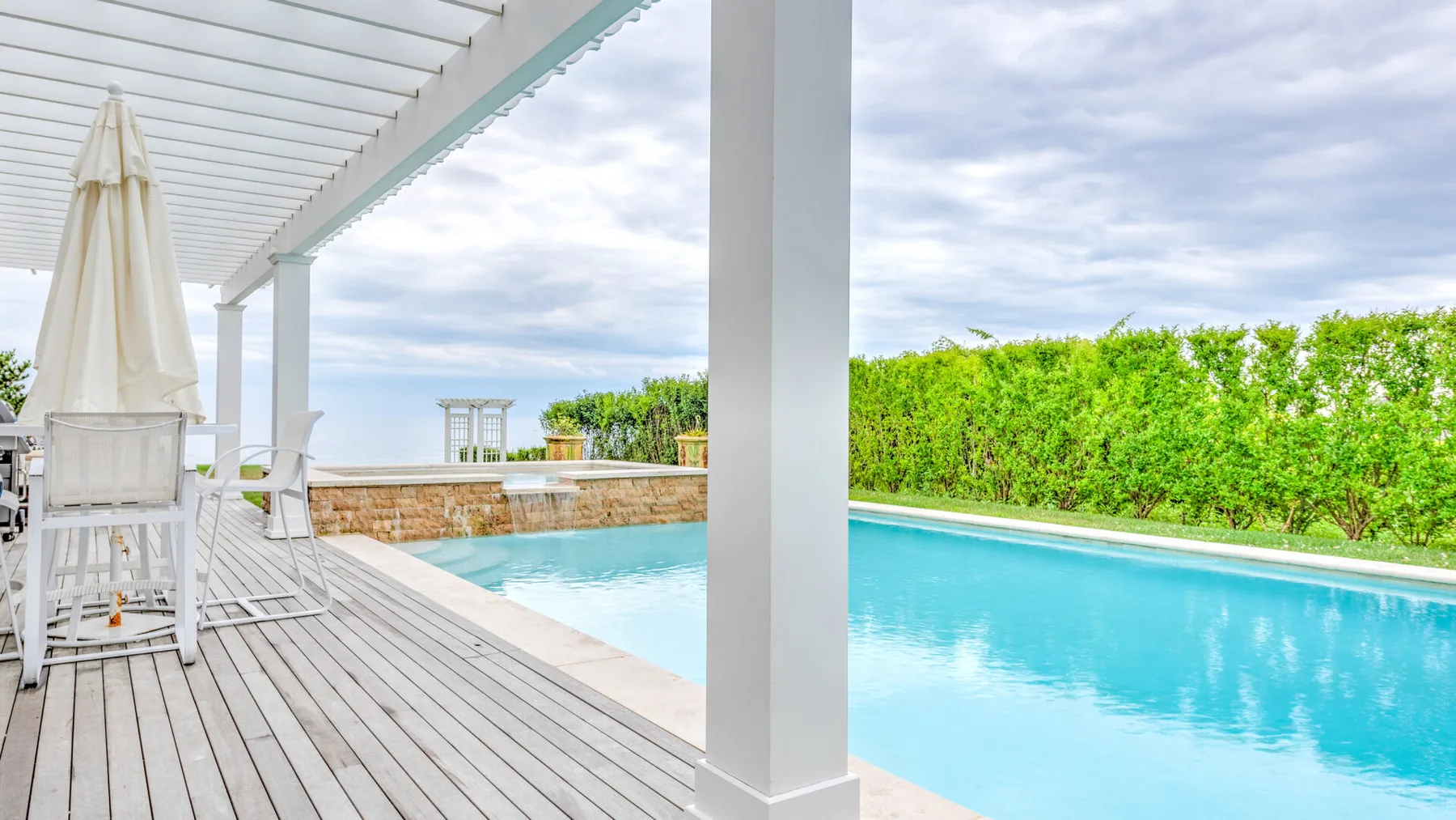 A covered outdoor patio opens onto a swimming pool beside a green hedge.