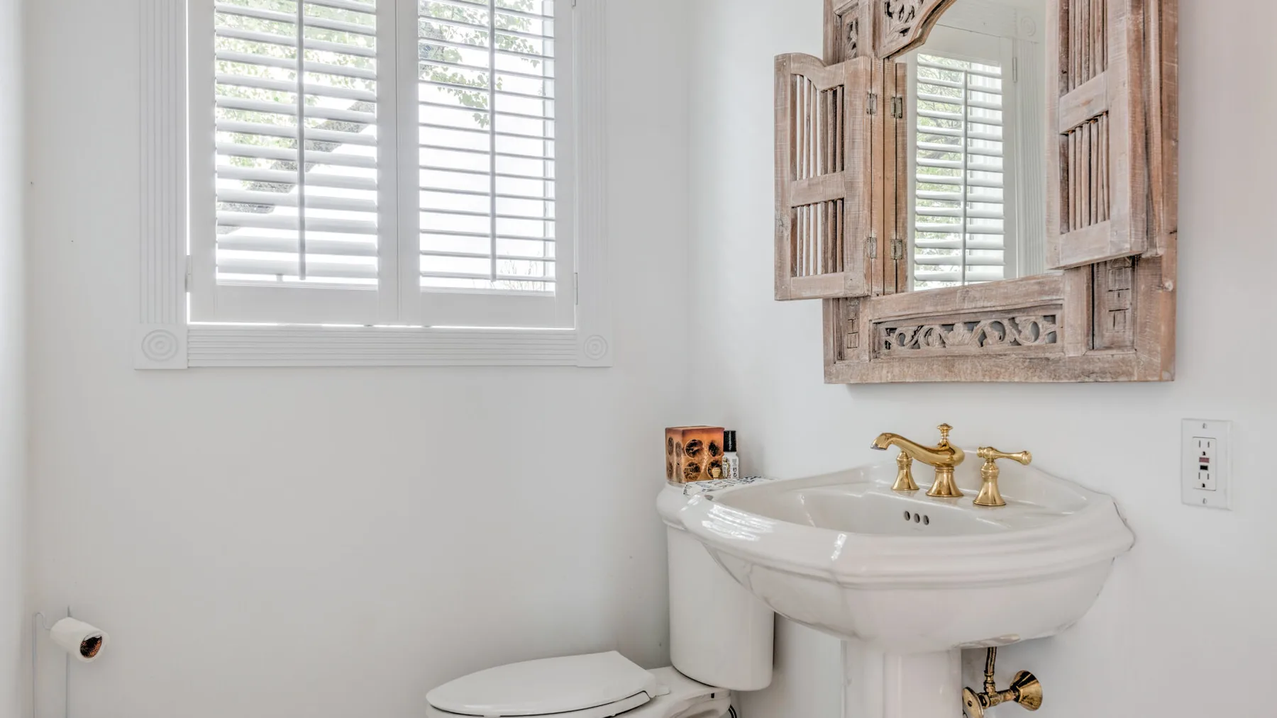 A bathroom with a white pedestal sink beneath a shuttered mirror.