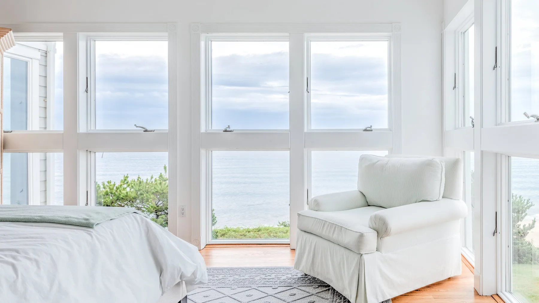 A bright bedroom with a bed and an armchair beside large windows overlooking the water.