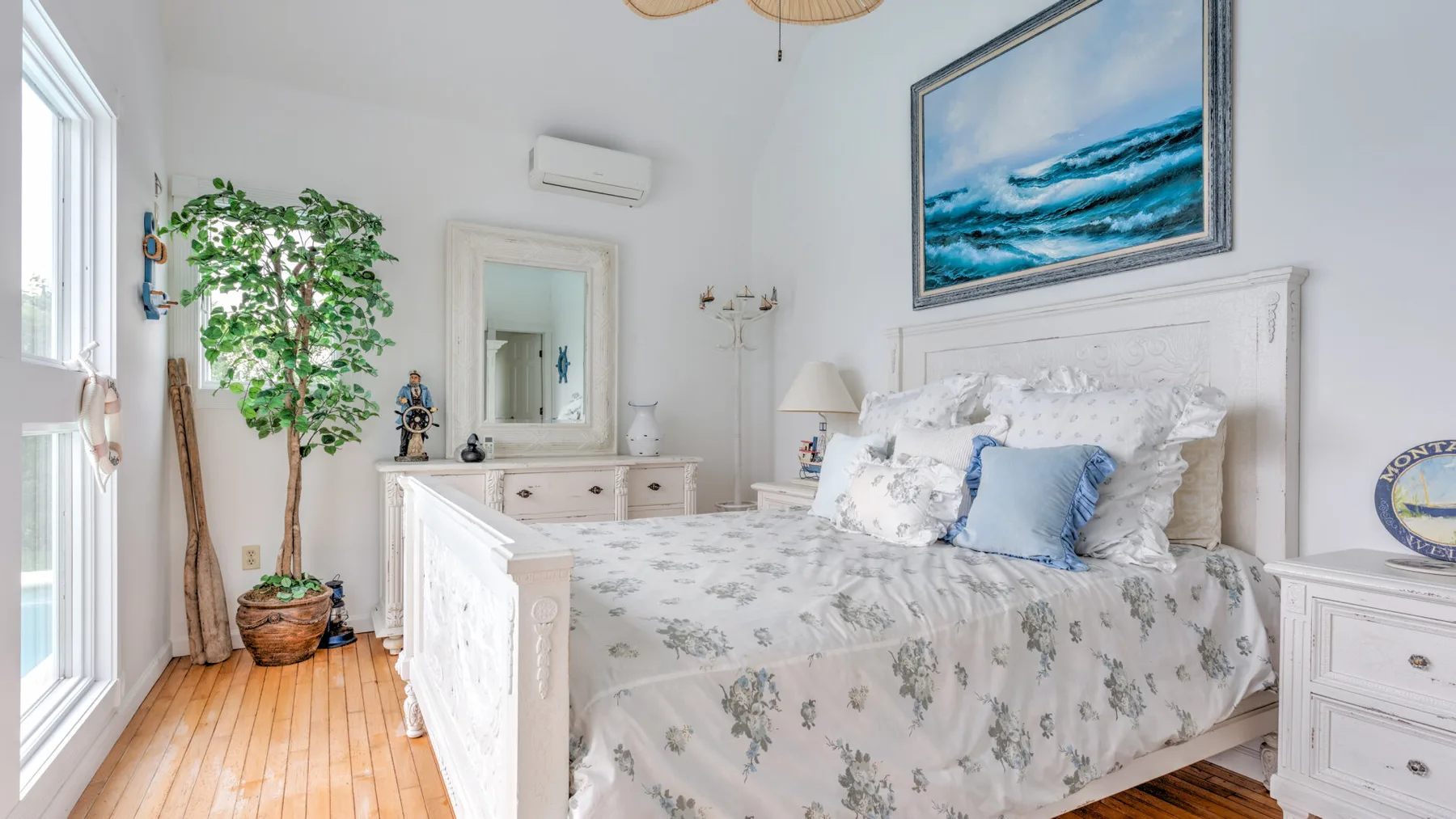 A bright bedroom with a floral bedspread, framed ocean artwork, and a potted plant.