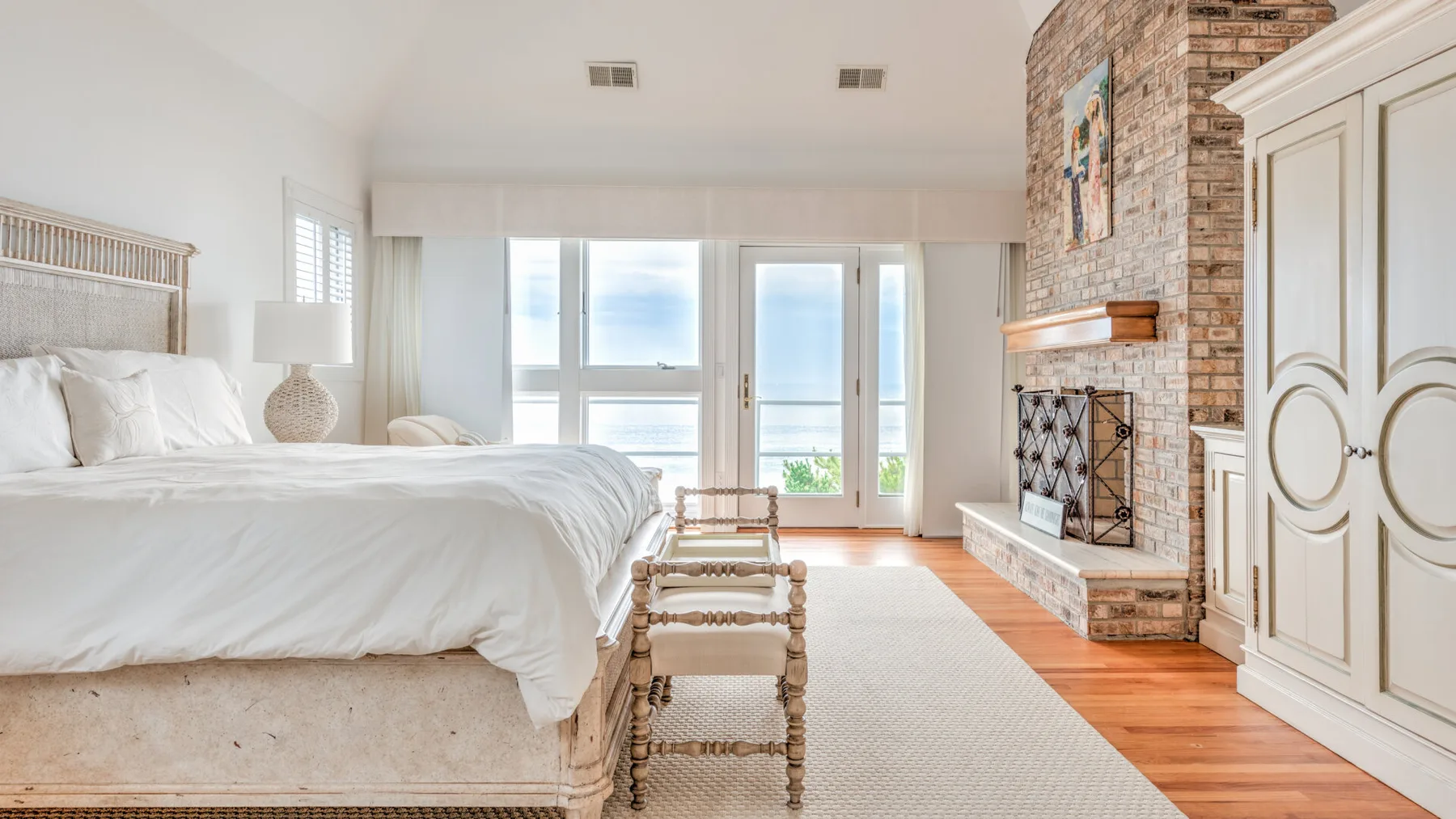 A bright bedroom with a white bed, large windows, and a brick fireplace.