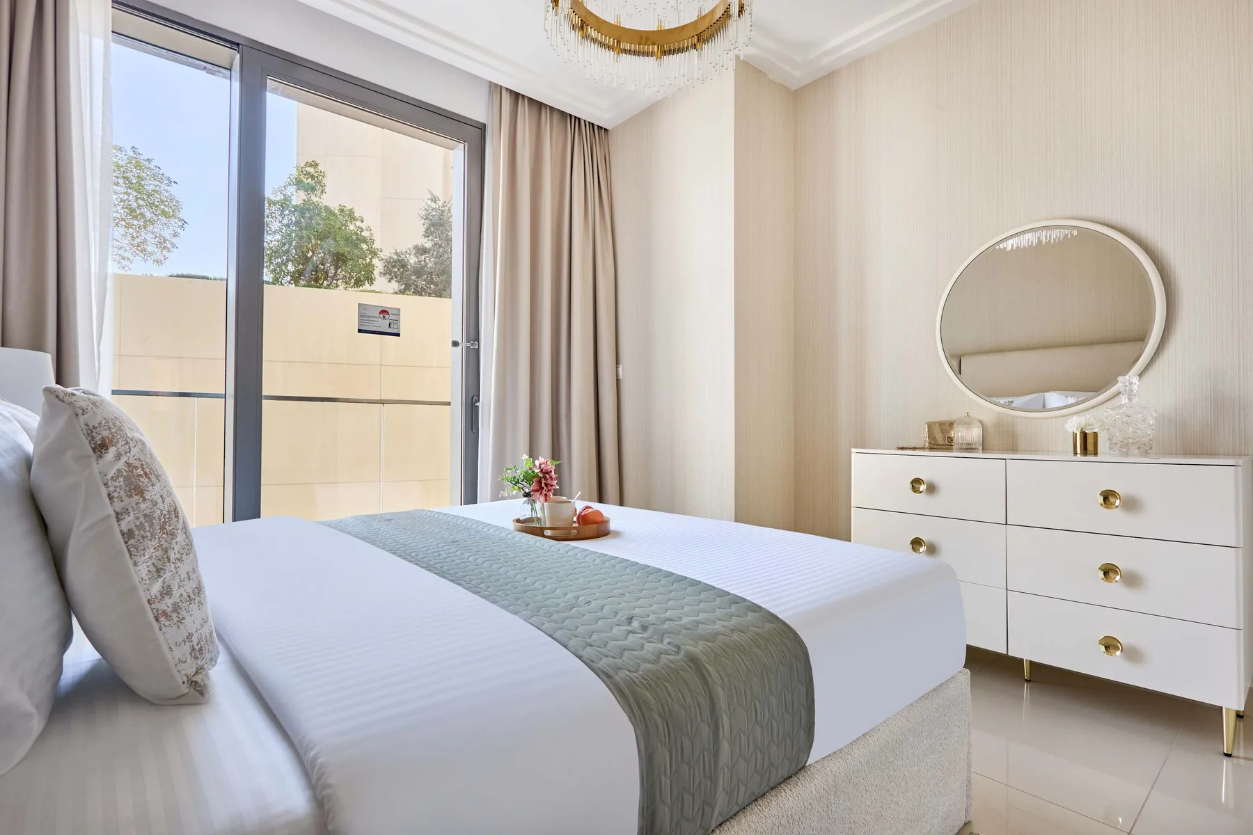 Un dormitorio luminoso con una cama bien hecha y una cómoda bajo un espejo redondo.