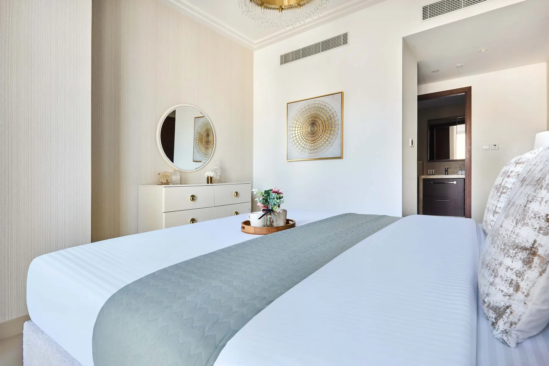 Un dormitorio luminoso con una cama tendida y una cómoda con un espejo redondo.