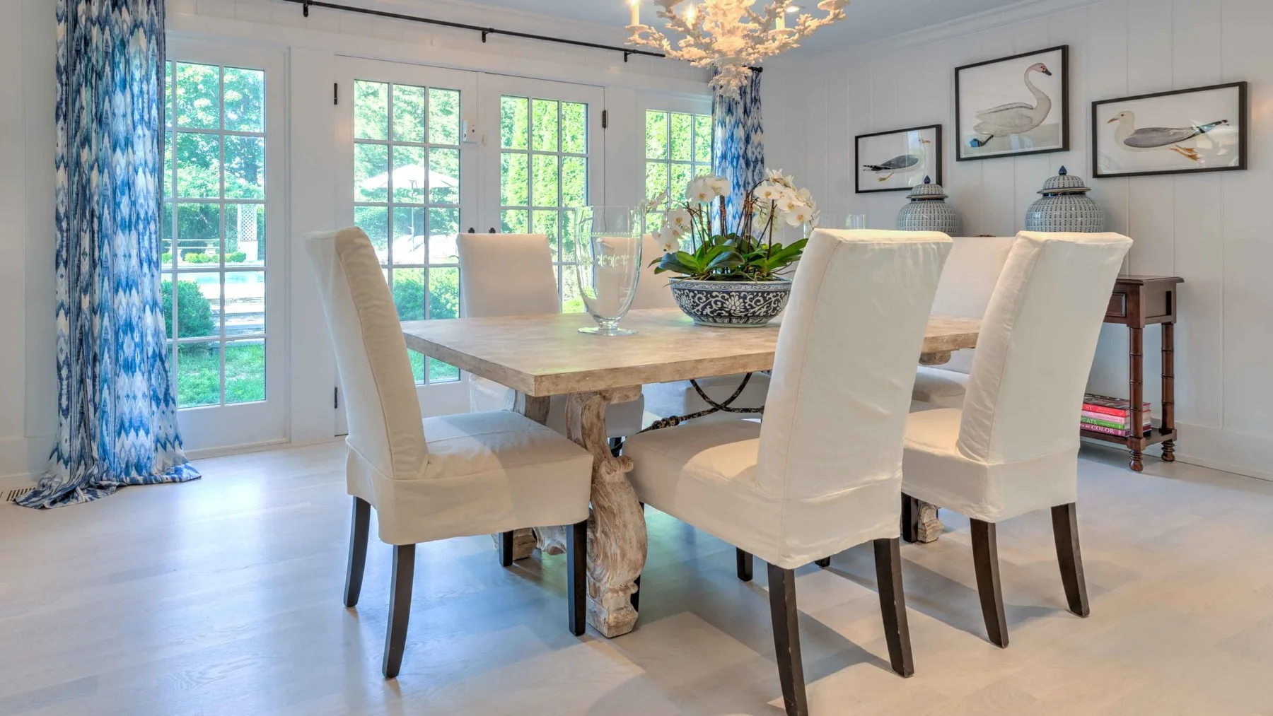 A bright dining room has a long table, upholstered seating, and glass doors opening to a garden.
