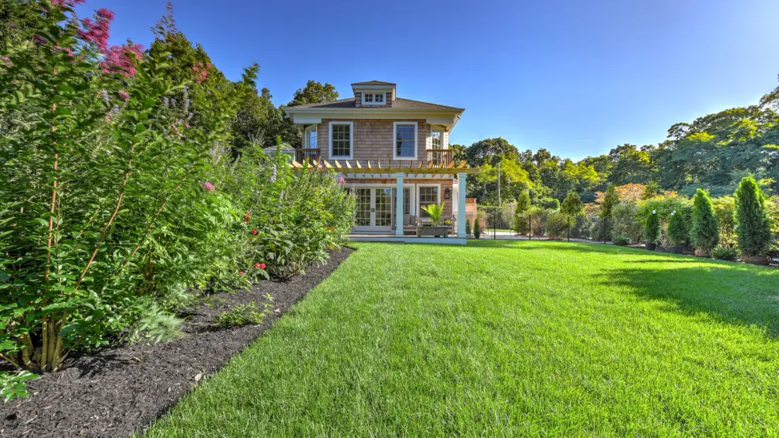 A house stands beyond a broad green lawn, surrounded by shrubs and trees.