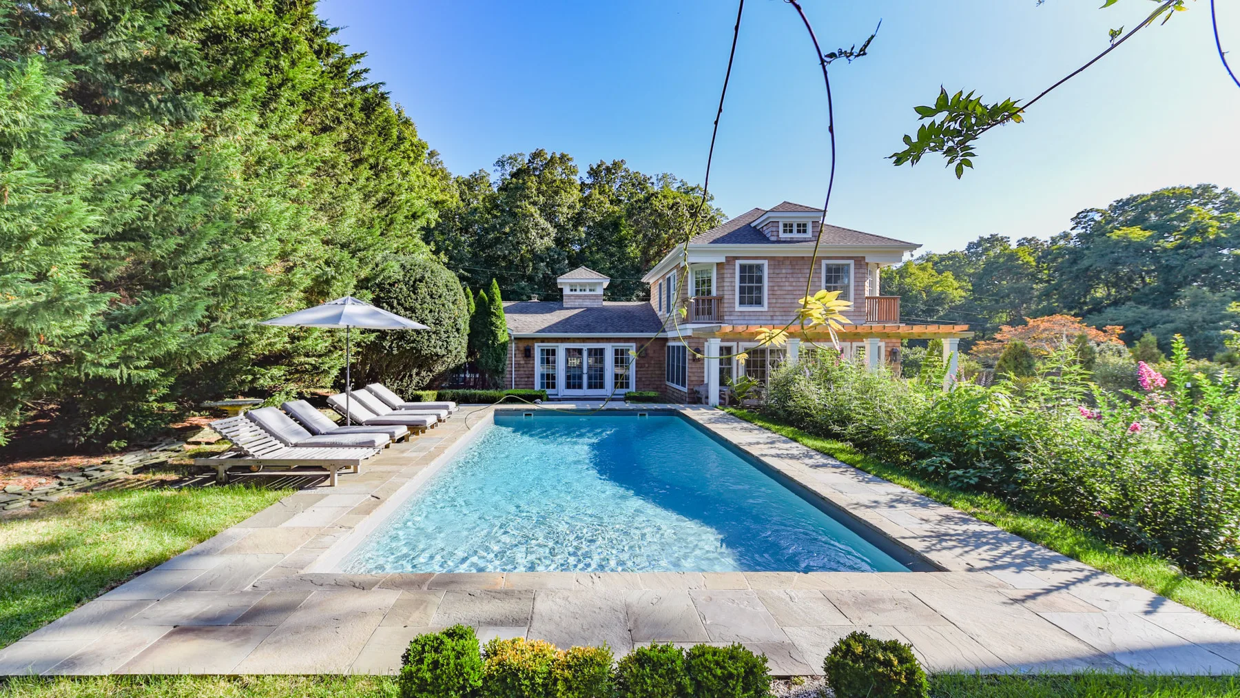 An outdoor swimming pool sits beside a landscaped garden and a large house.