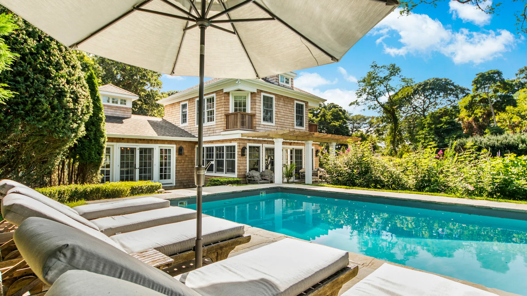 An outdoor swimming pool beside a house, with lounge seating under a large umbrella.