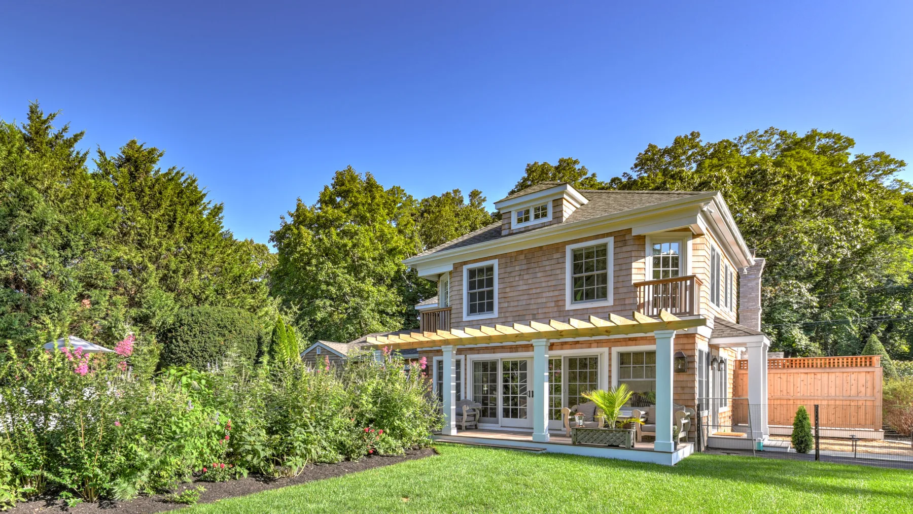 A two-story house stands behind a green lawn and leafy garden.