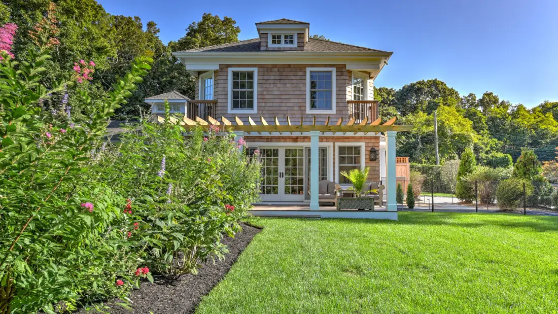 A two-story house with a wooden deck overlooks a green lawn and garden.