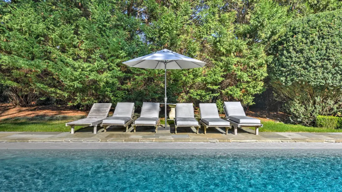 Outdoor swimming pool with lounge chairs and an umbrella beside a hedge.