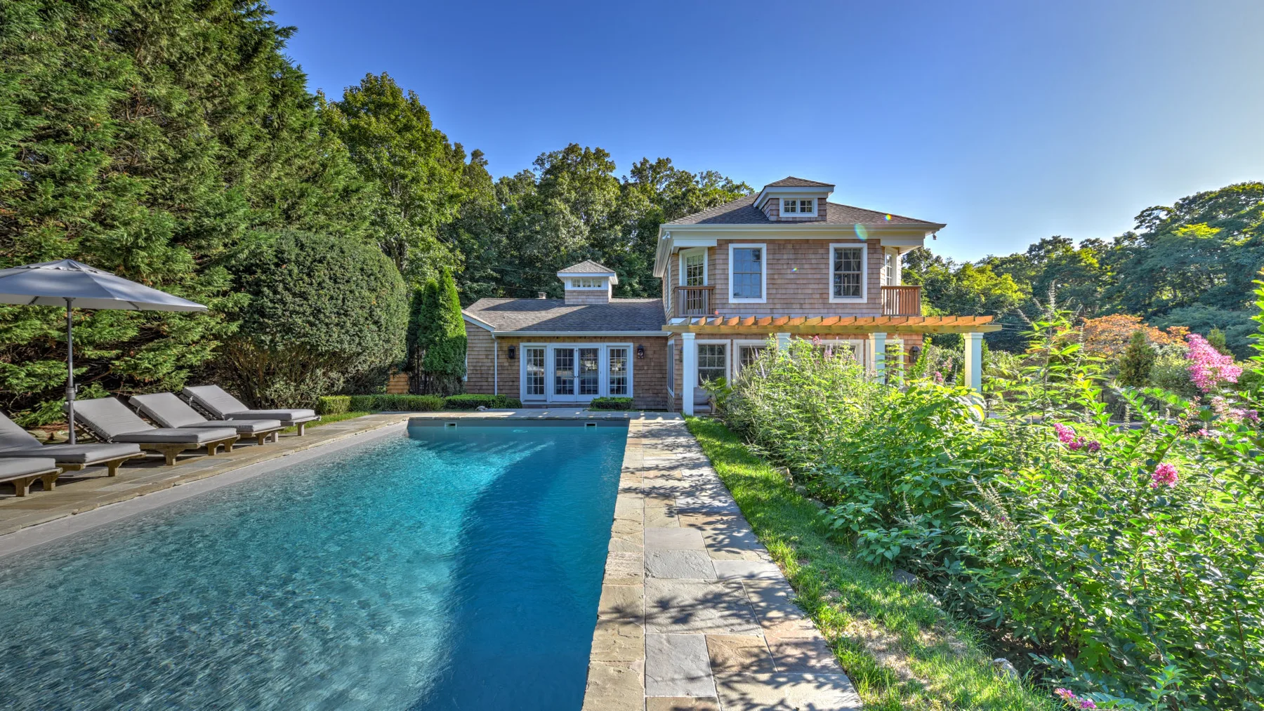 An outdoor swimming pool sits beside a large house and leafy garden.