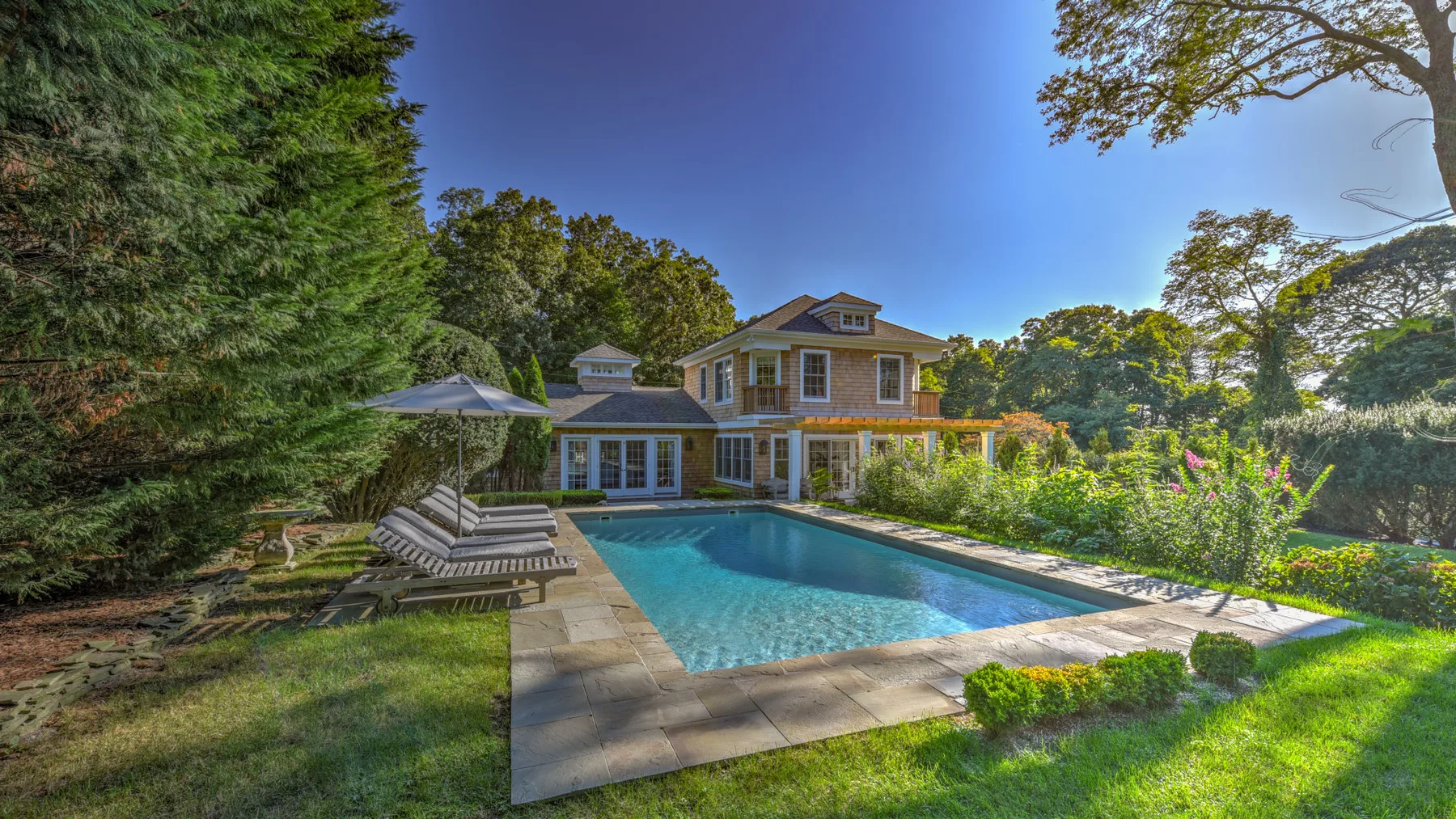 A swimming pool beside a large house, surrounded by trees and gardens.
