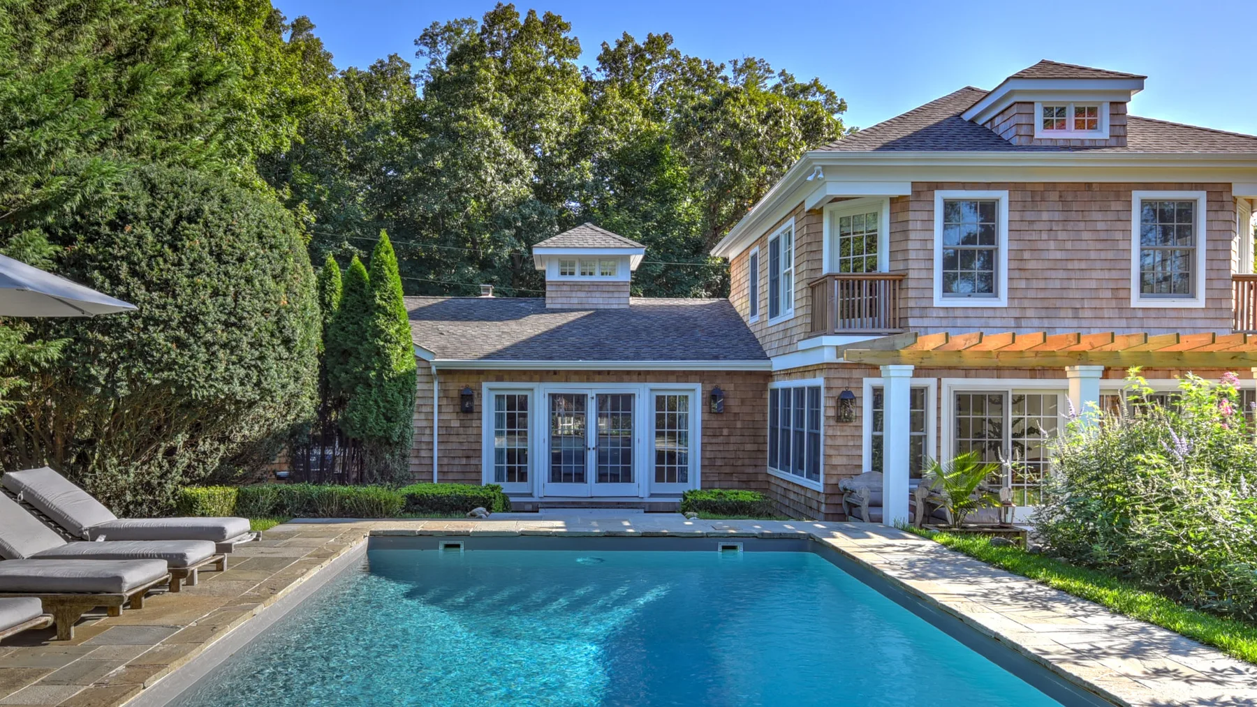 A swimming pool sits beside a large house, surrounded by trees and greenery.