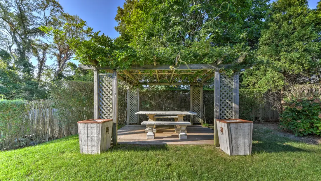 An outdoor picnic table sits beneath a leafy pergola in a garden.