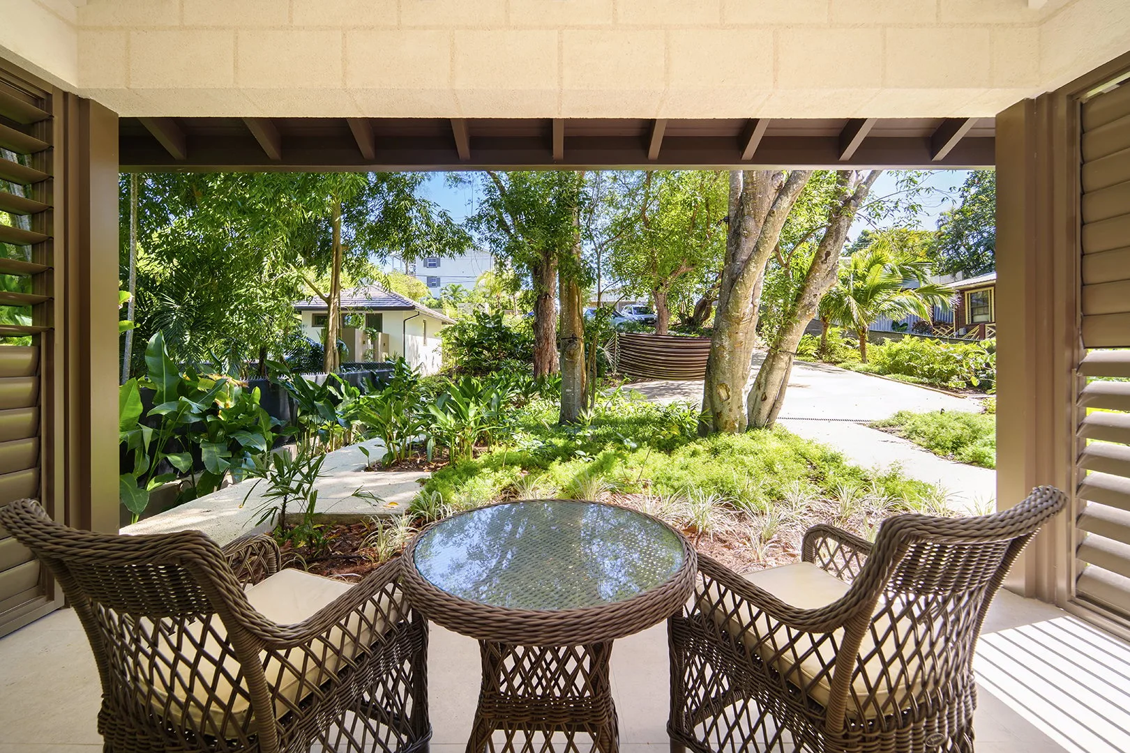 A covered patio with woven seating and a round glass-top table overlooks a garden.