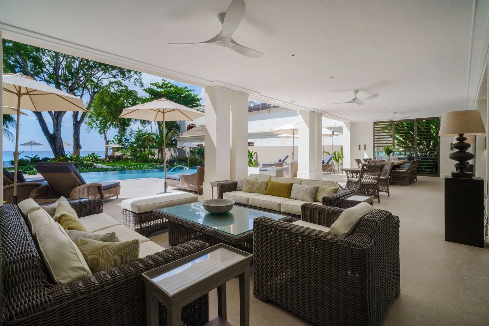 A covered outdoor lounge with cushioned seating overlooks a pool and landscaped grounds.