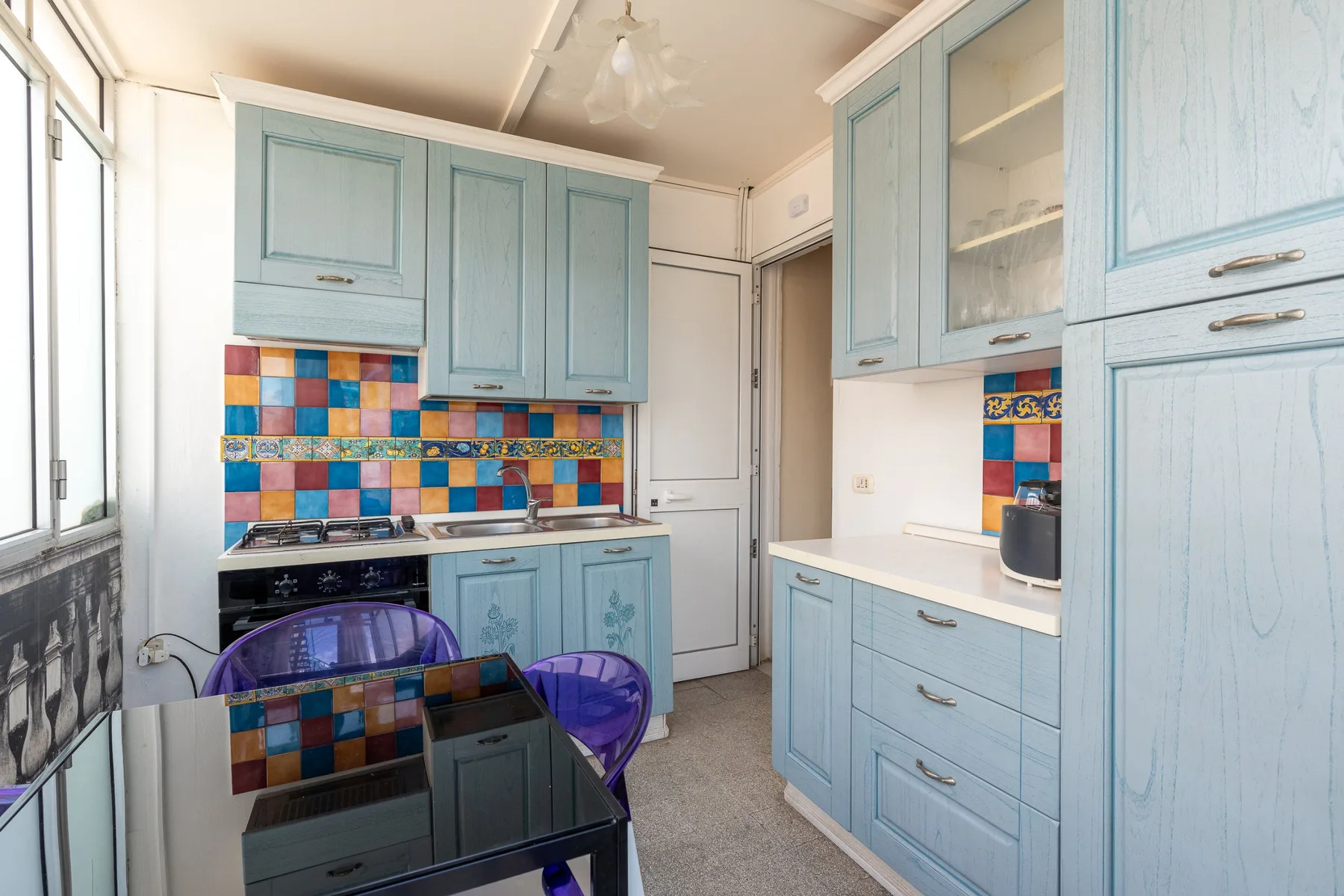 Una cocina luminosa con gabinetes azul claro, azulejos coloridos y una mesa de vidrio.