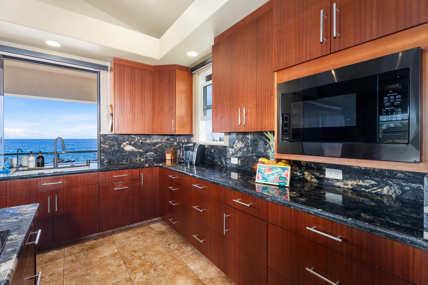 Una cocina con gabinetes de madera rojiza, encimeras oscuras y una ventana con vista al agua.