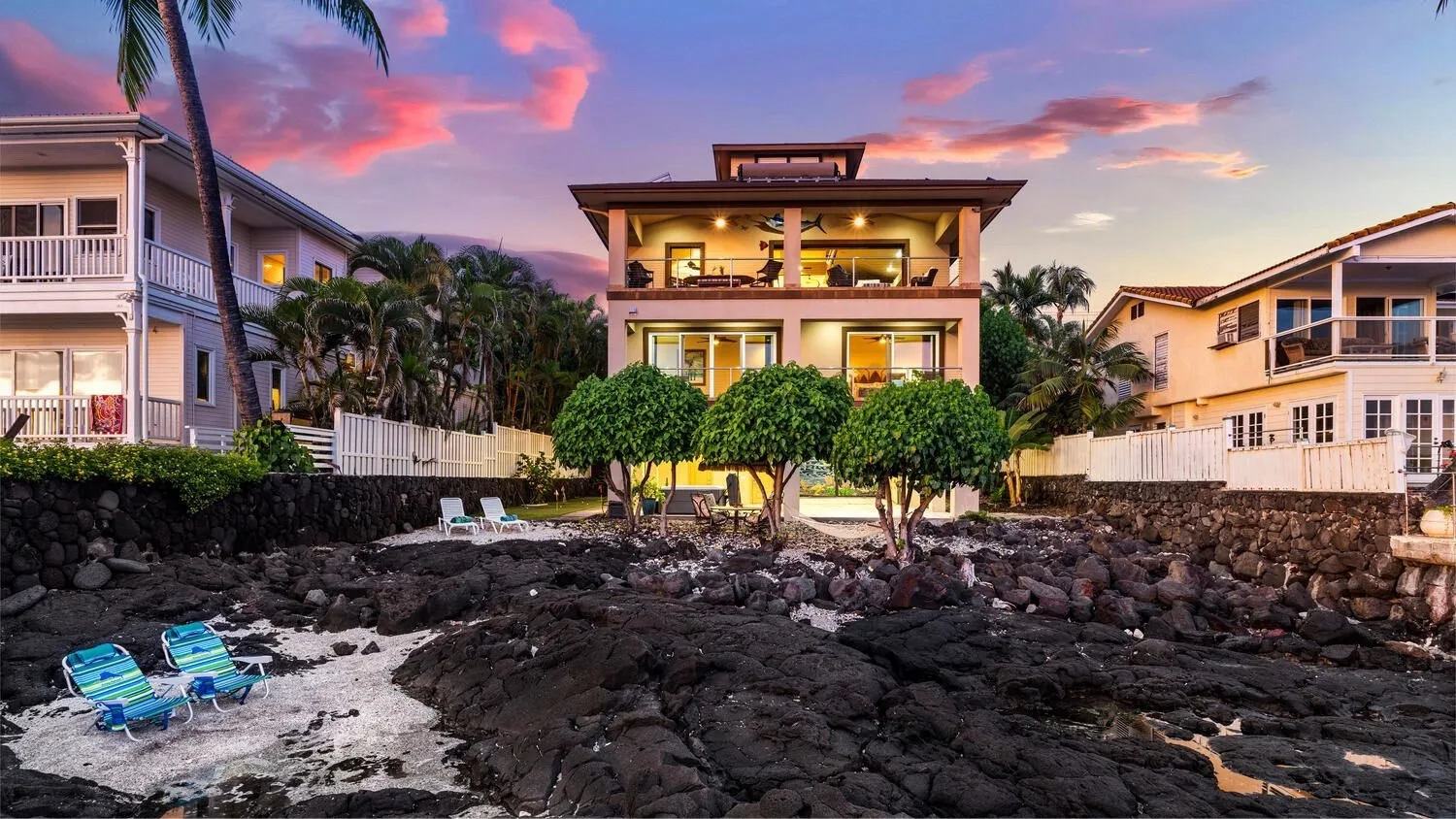 Una casa junto al agua se alza tras una costa rocosa bajo un cielo colorido al atardecer.
