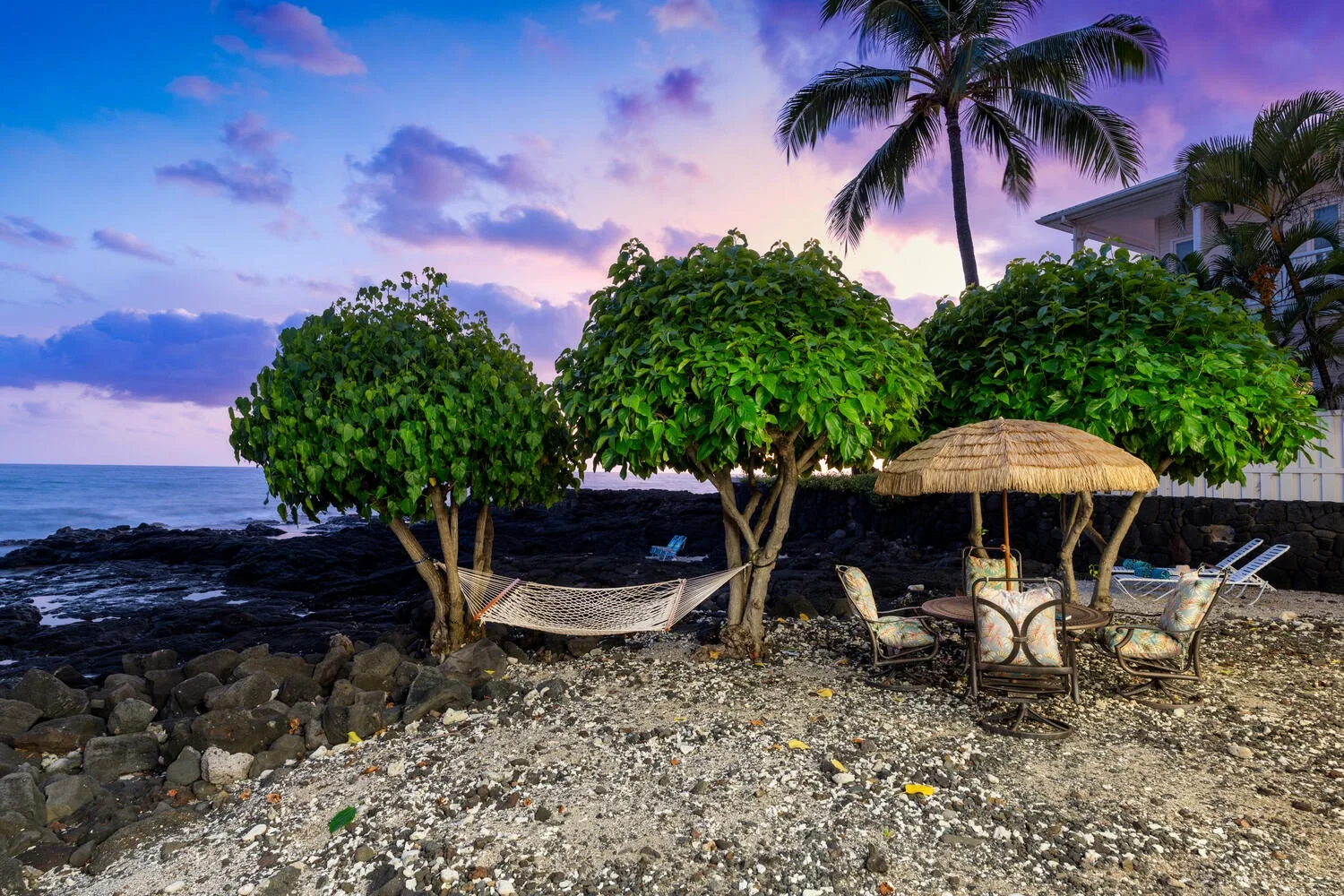 Una zona de descanso junto a la costa tropical, con árboles, una cubierta de paja y vistas al mar.