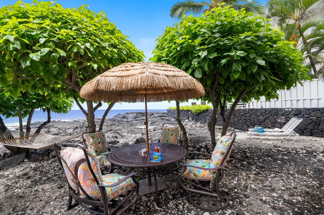 Una zona de asientos al aire libre bajo una sombrilla de paja, rodeada de árboles frondosos.
