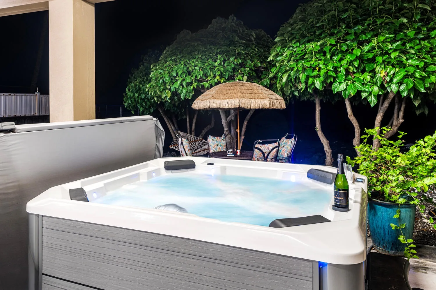 Una bañera de hidromasaje exterior está junto a árboles frondosos y asientos de patio por la noche.