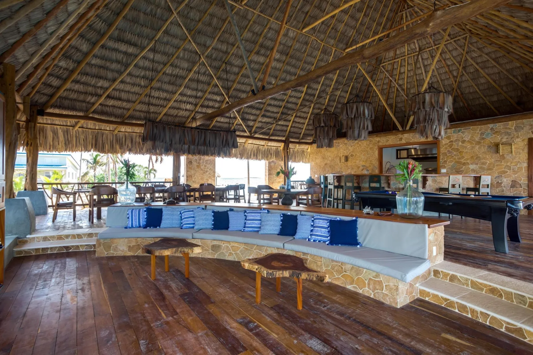 An open-air lounge with a long cushioned seating area beneath a thatched roof.