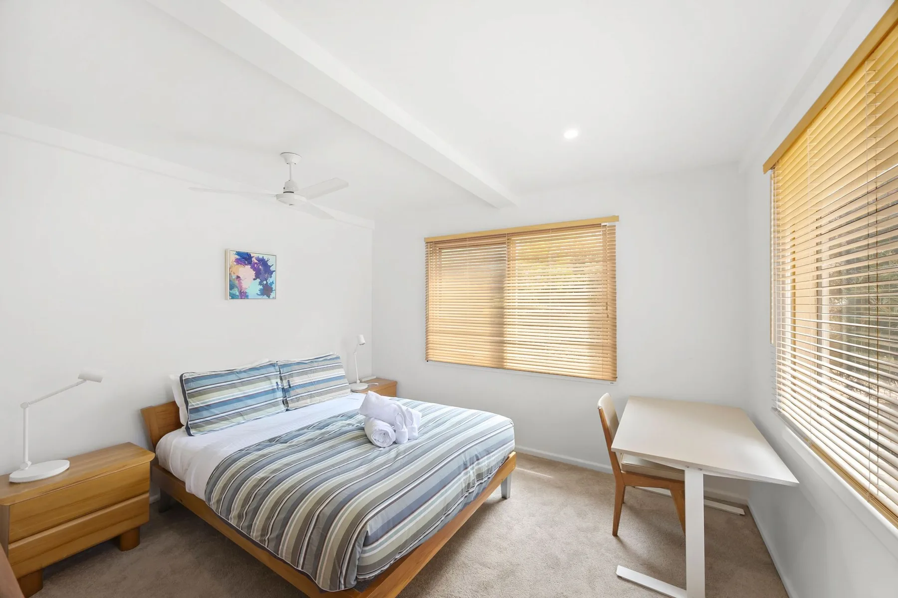 A bright bedroom with a striped bed and a small desk by the windows.
