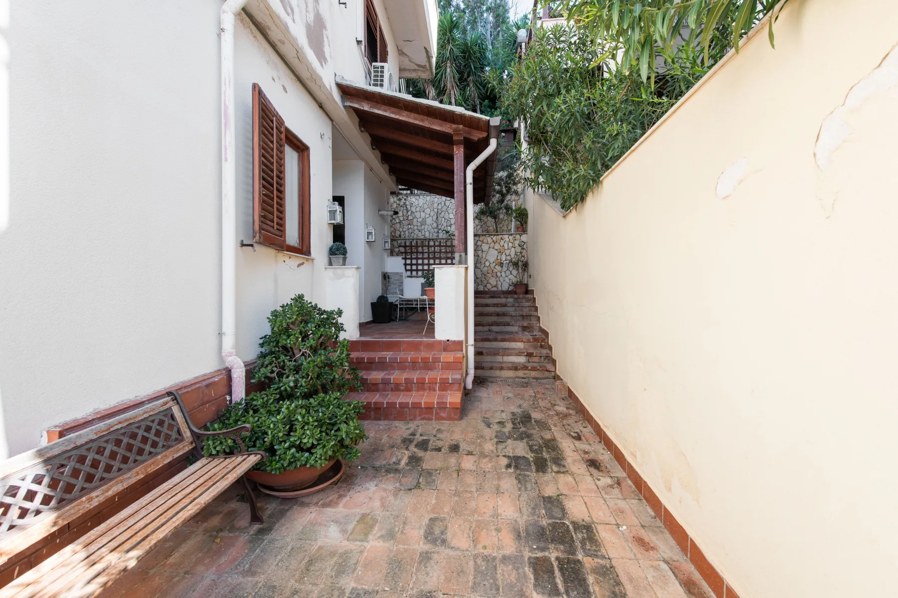 A paved outdoor passage runs between a building and a high wall, with steps at the far end.