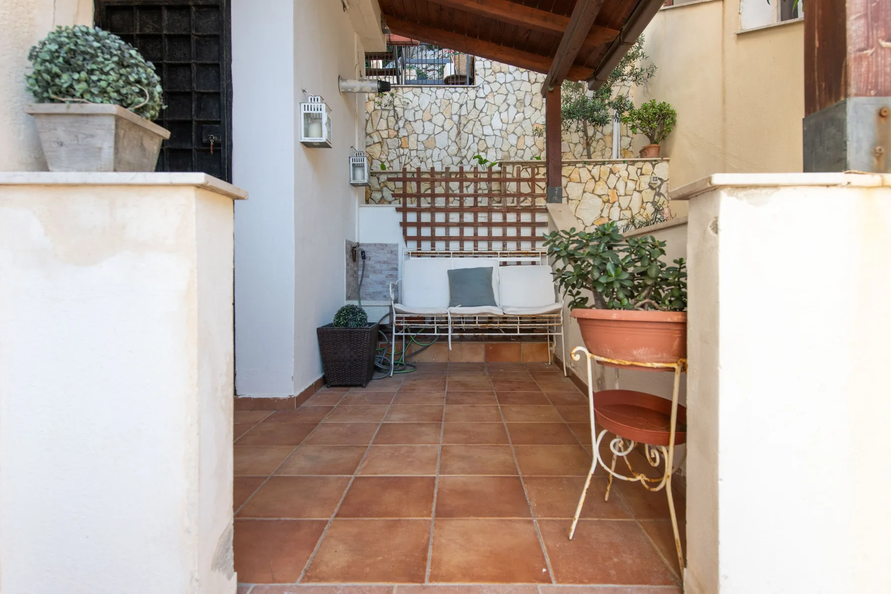 A covered patio with seating and potted plants along a stone wall.