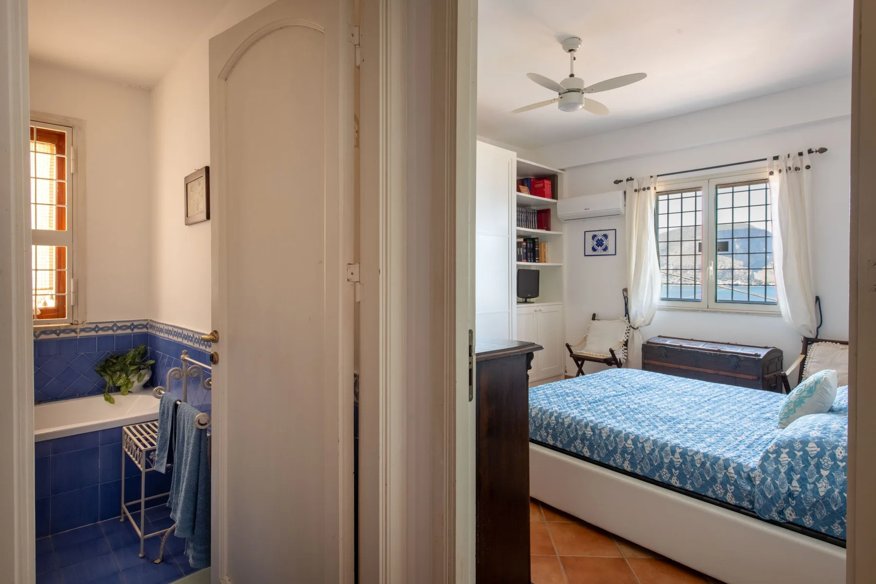 A bedroom with a blue patterned bedspread, seen through an open doorway beside a bathroom.