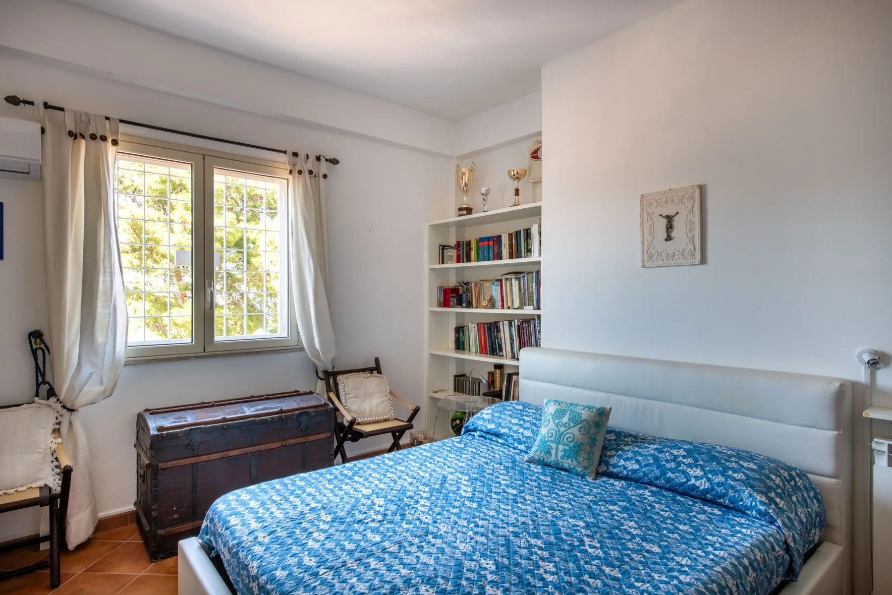 A bedroom with a blue patterned bed beside a window and bookshelves.