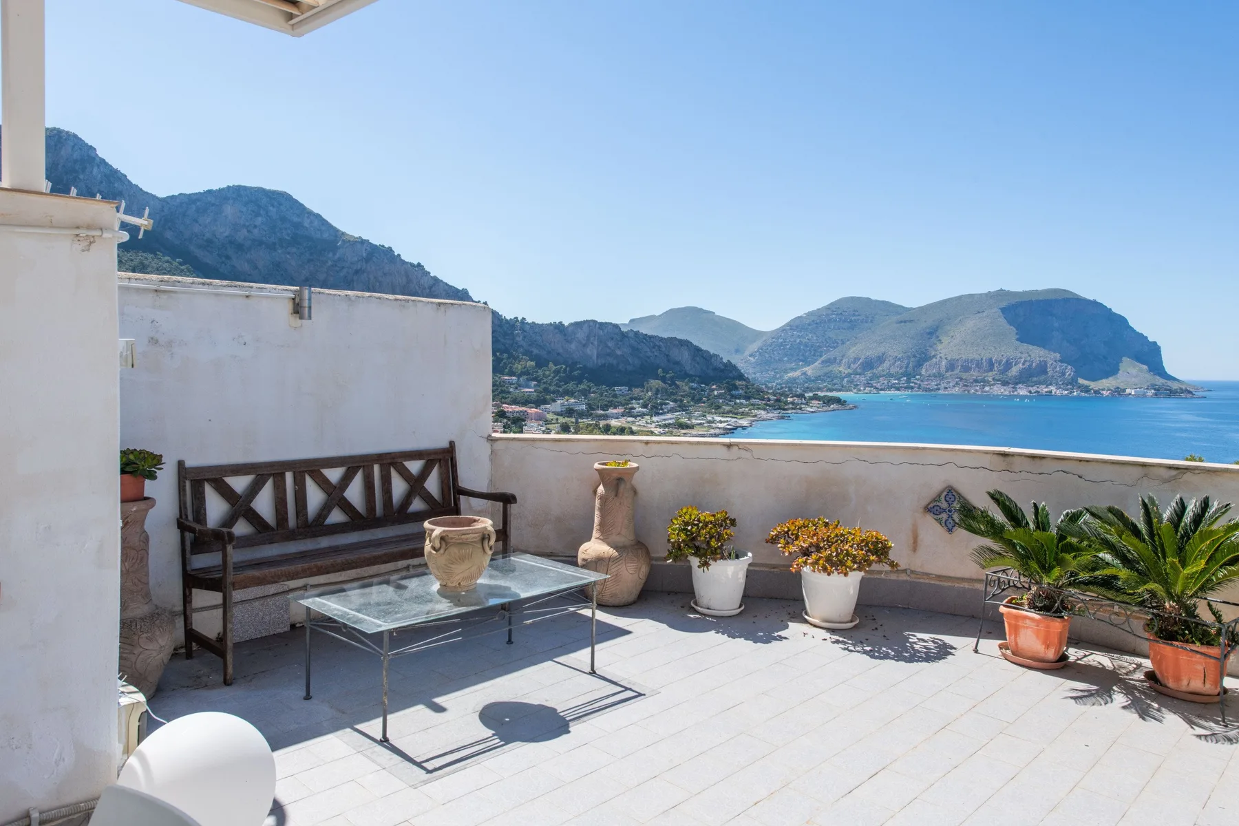 A sunlit terrace overlooks the sea, with outdoor seating and potted plants.