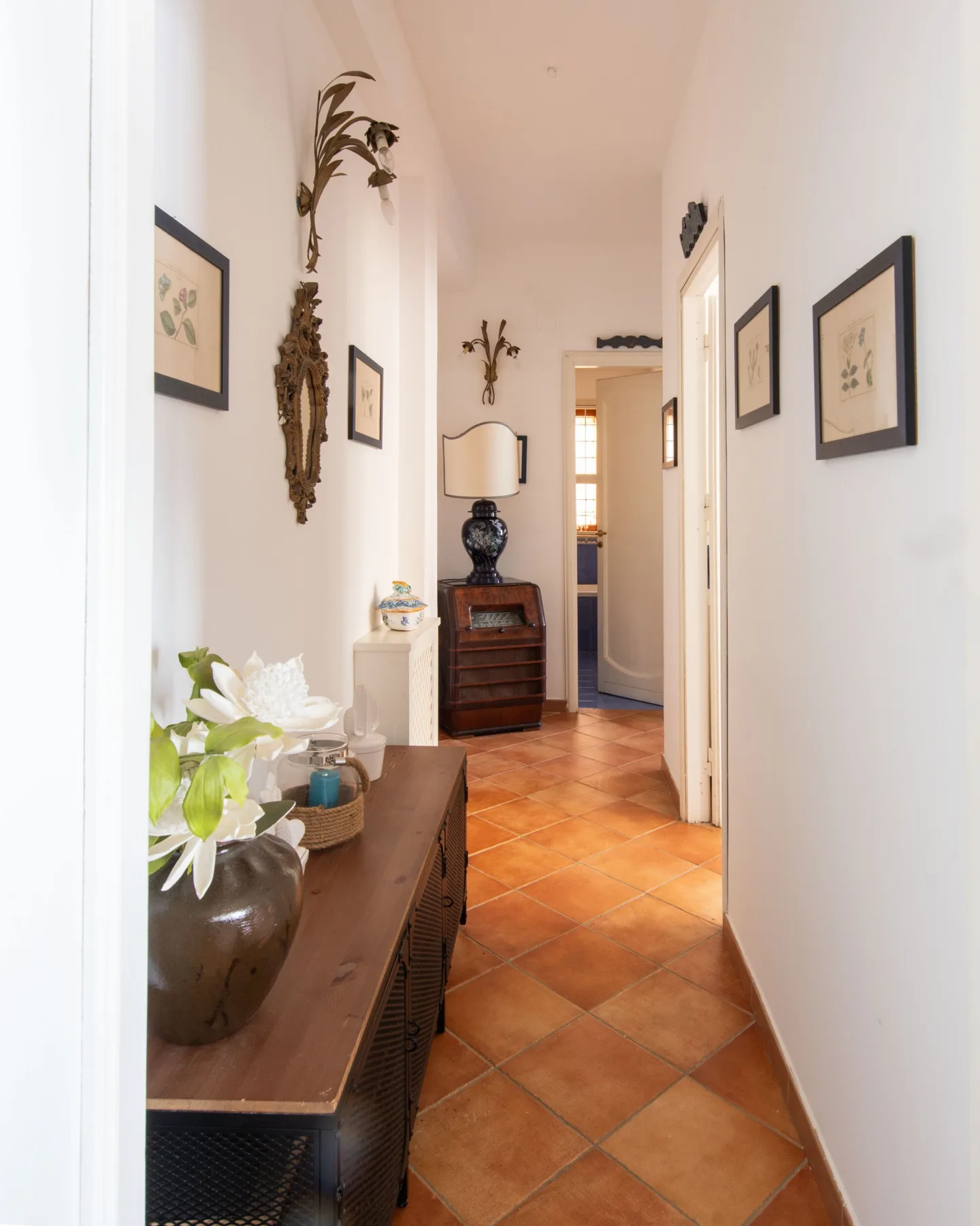 A narrow hallway with terracotta-colored floor tiles and framed pictures along the walls.