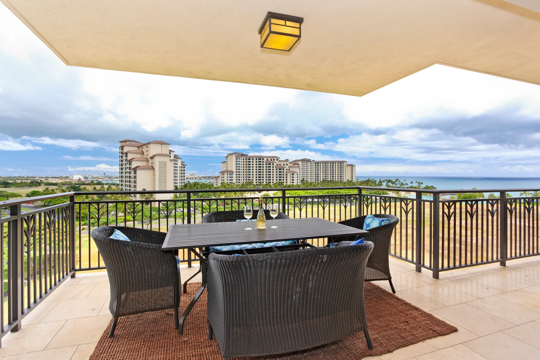 A covered balcony has a dining table and seating overlooking nearby buildings and greenery.