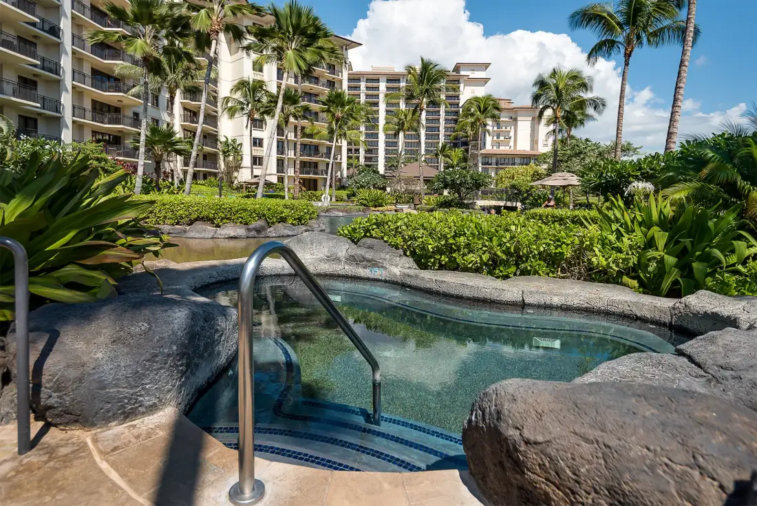A small outdoor pool sits among tropical landscaping in front of multi-story buildings.