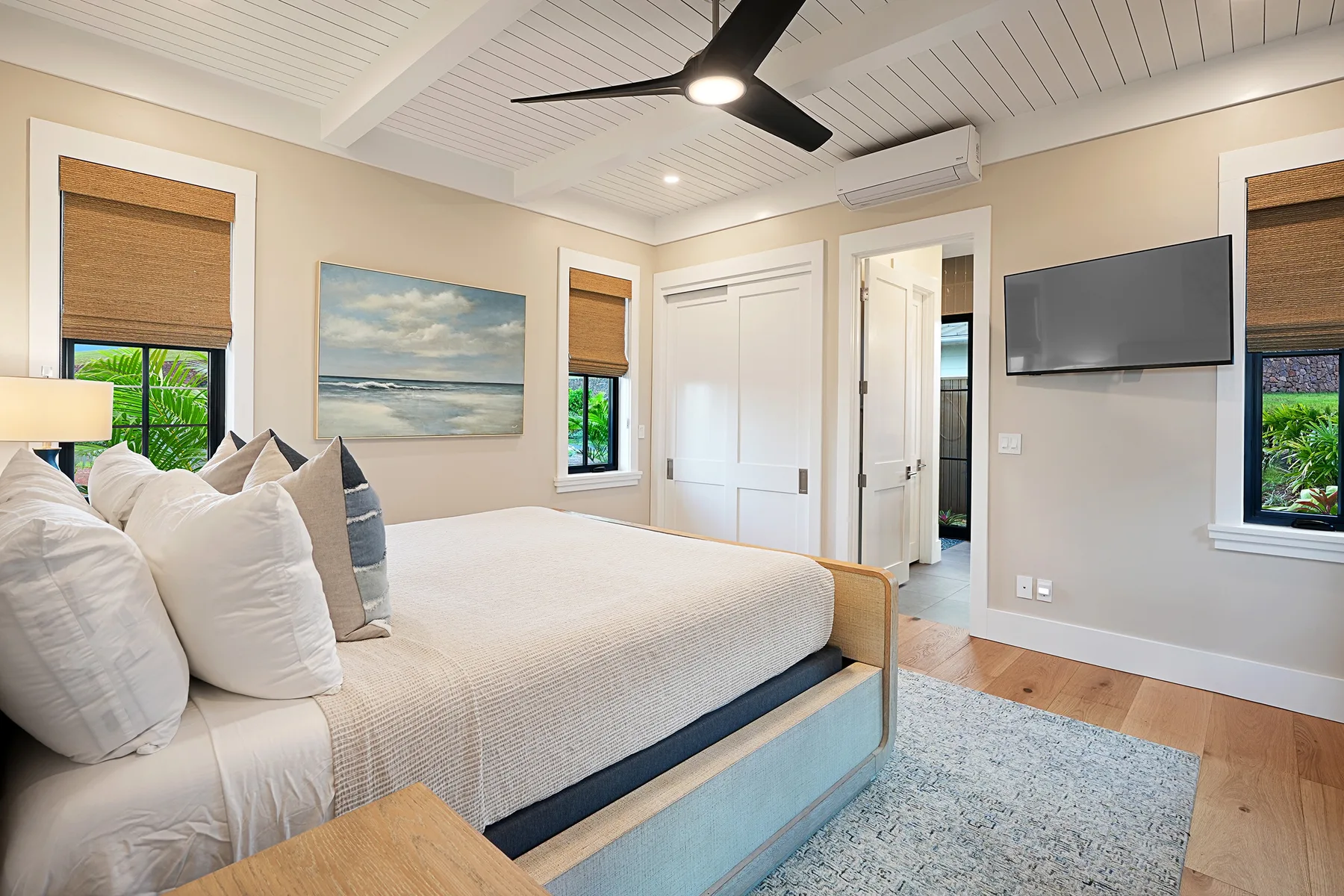A bright bedroom with a bed, wall-mounted television, and windows with woven shades.