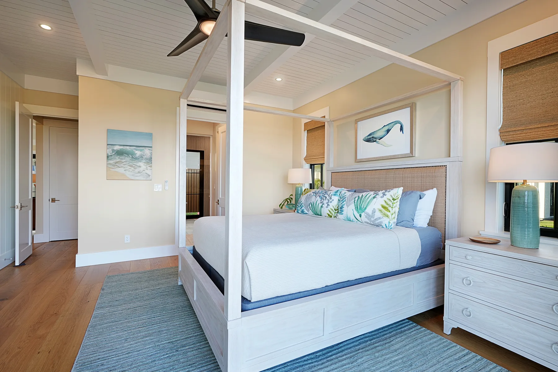 A bright bedroom features a canopy bed with patterned pillows and framed coastal artwork.