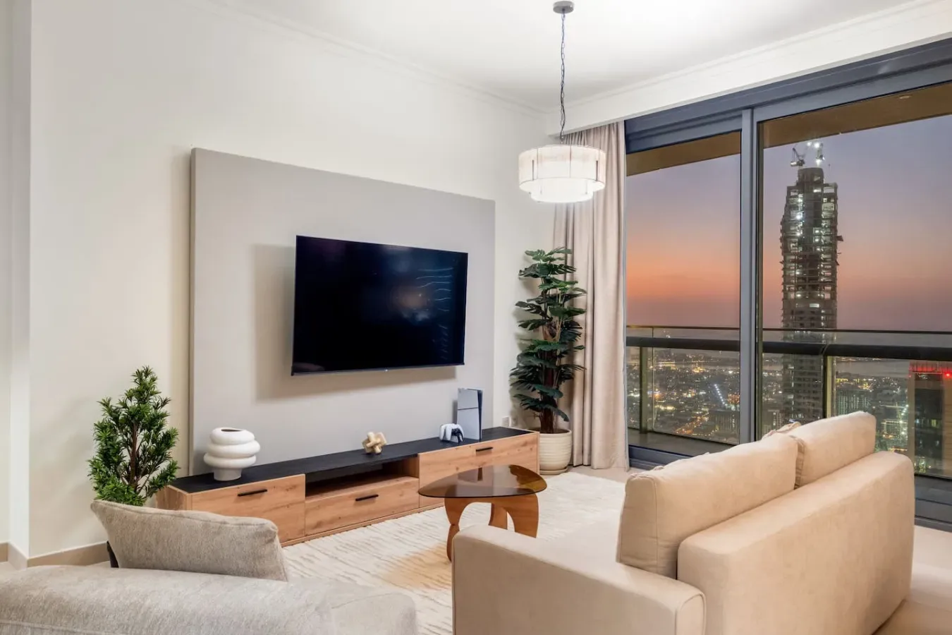 A living room with a television, seating, and floor-to-ceiling windows overlooking the city at sunset.