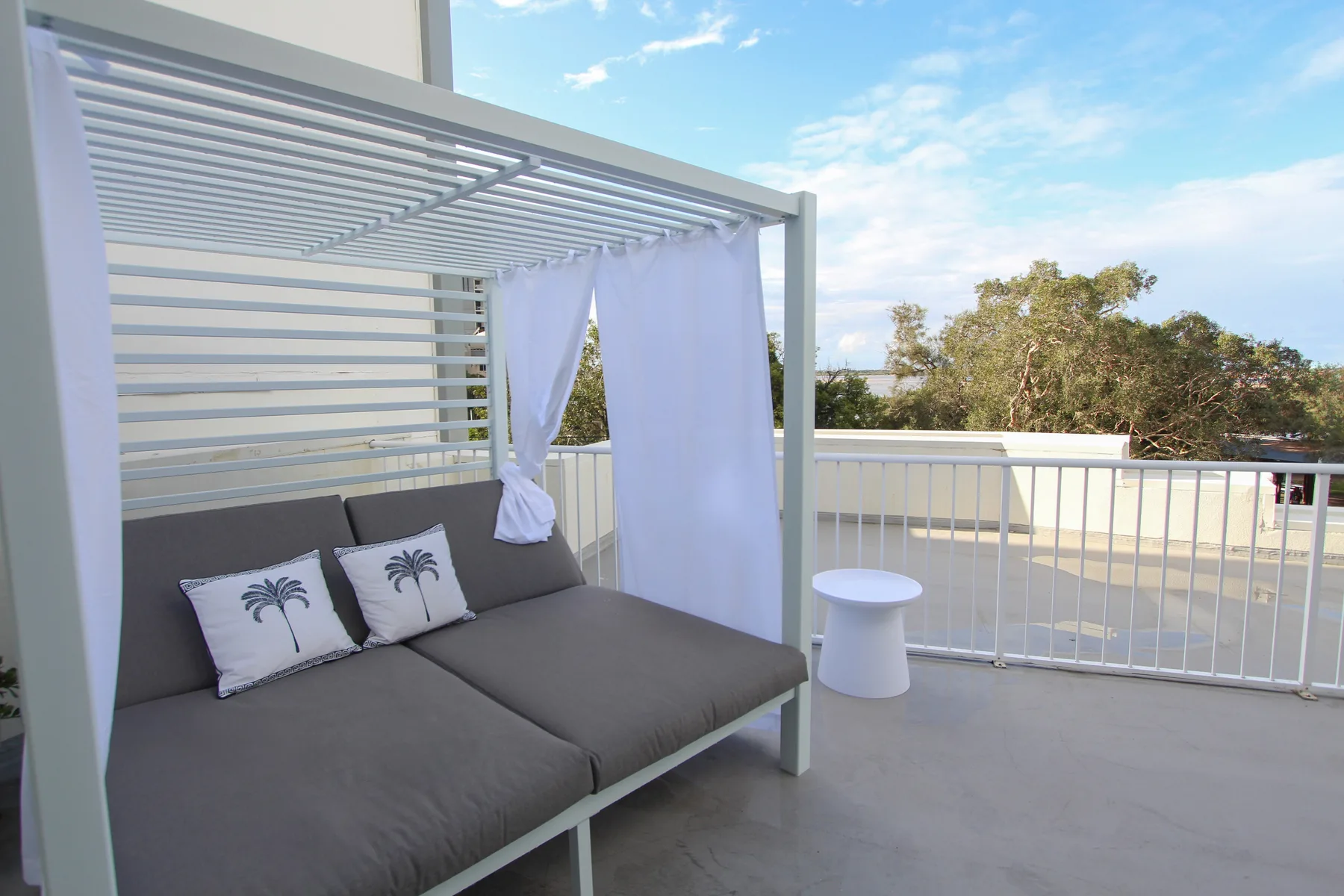 Una terraza exterior tiene un diván acolchado bajo una pérgola de listones.