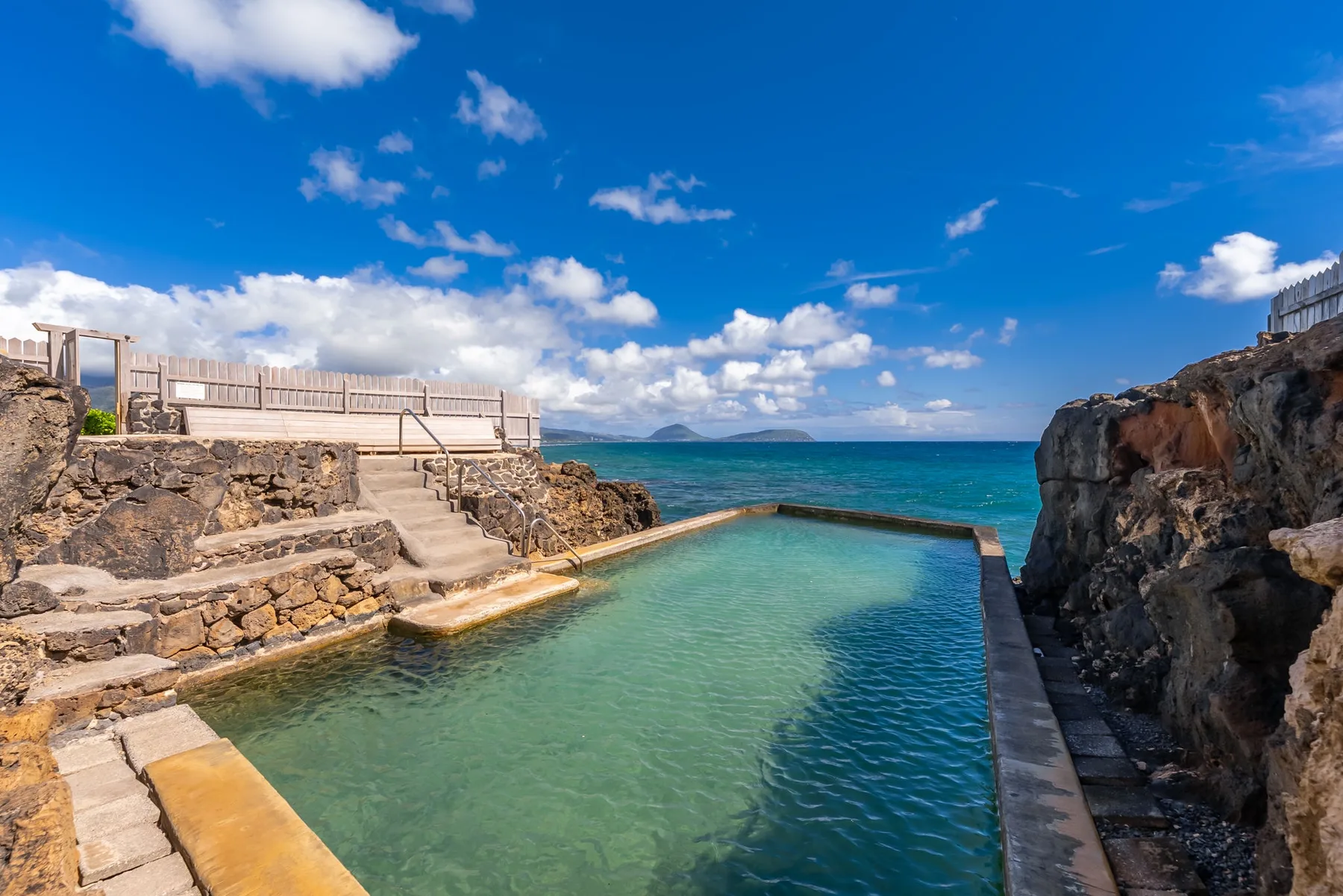A seaside swimming pool sits among rocky stone terraces, with the ocean beyond.