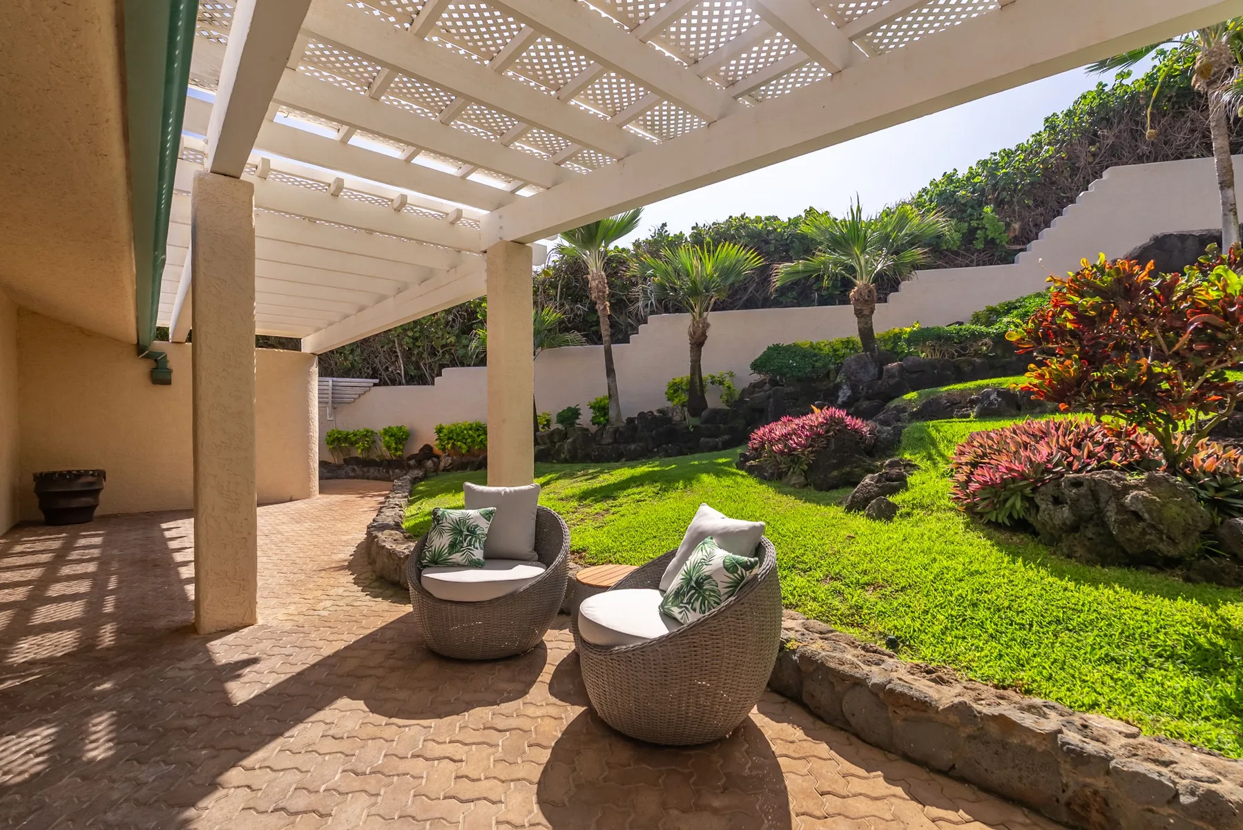A shaded patio with two outdoor seats overlooks a landscaped garden.