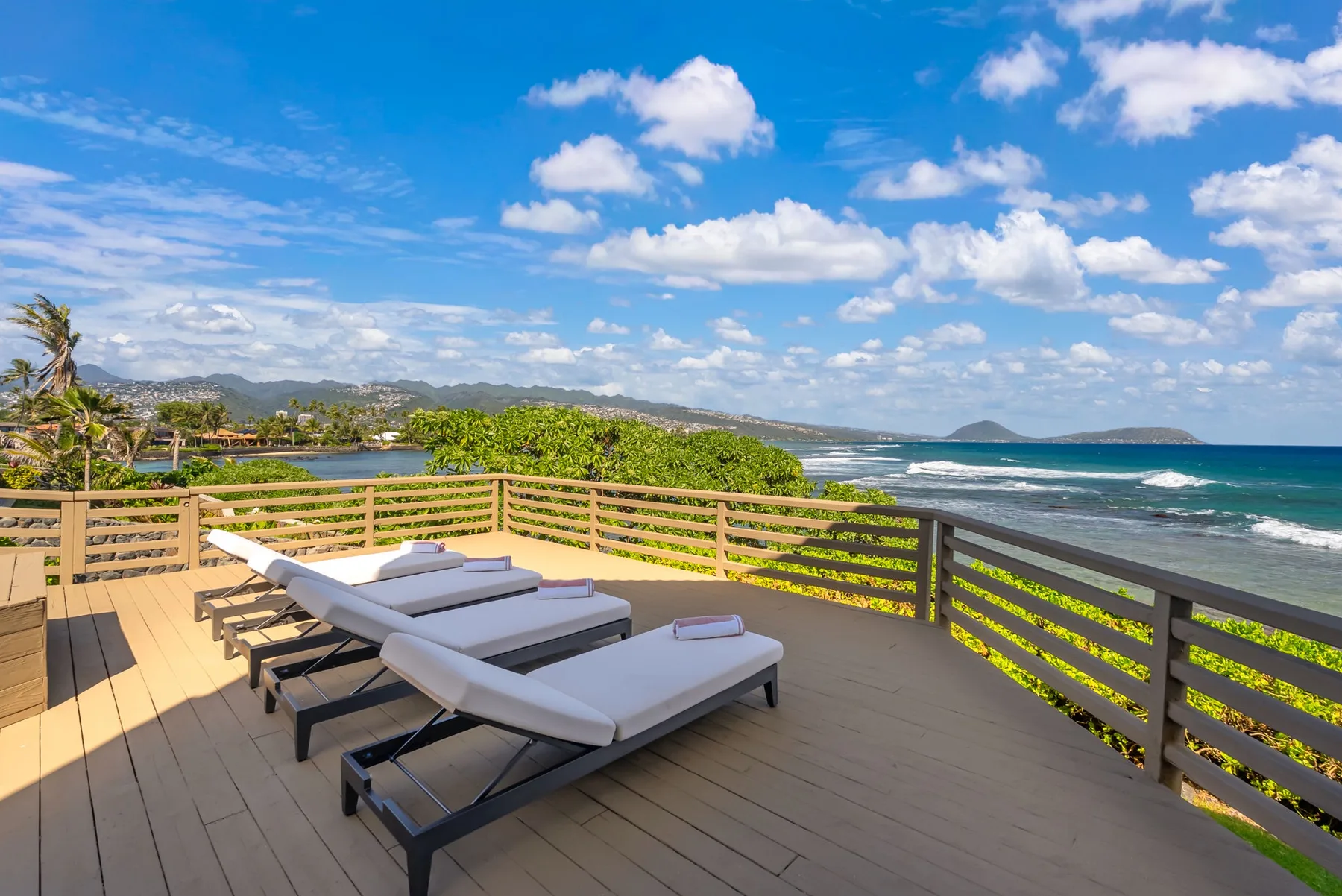 A wooden deck with lounge seating overlooks the ocean and a green coastline.