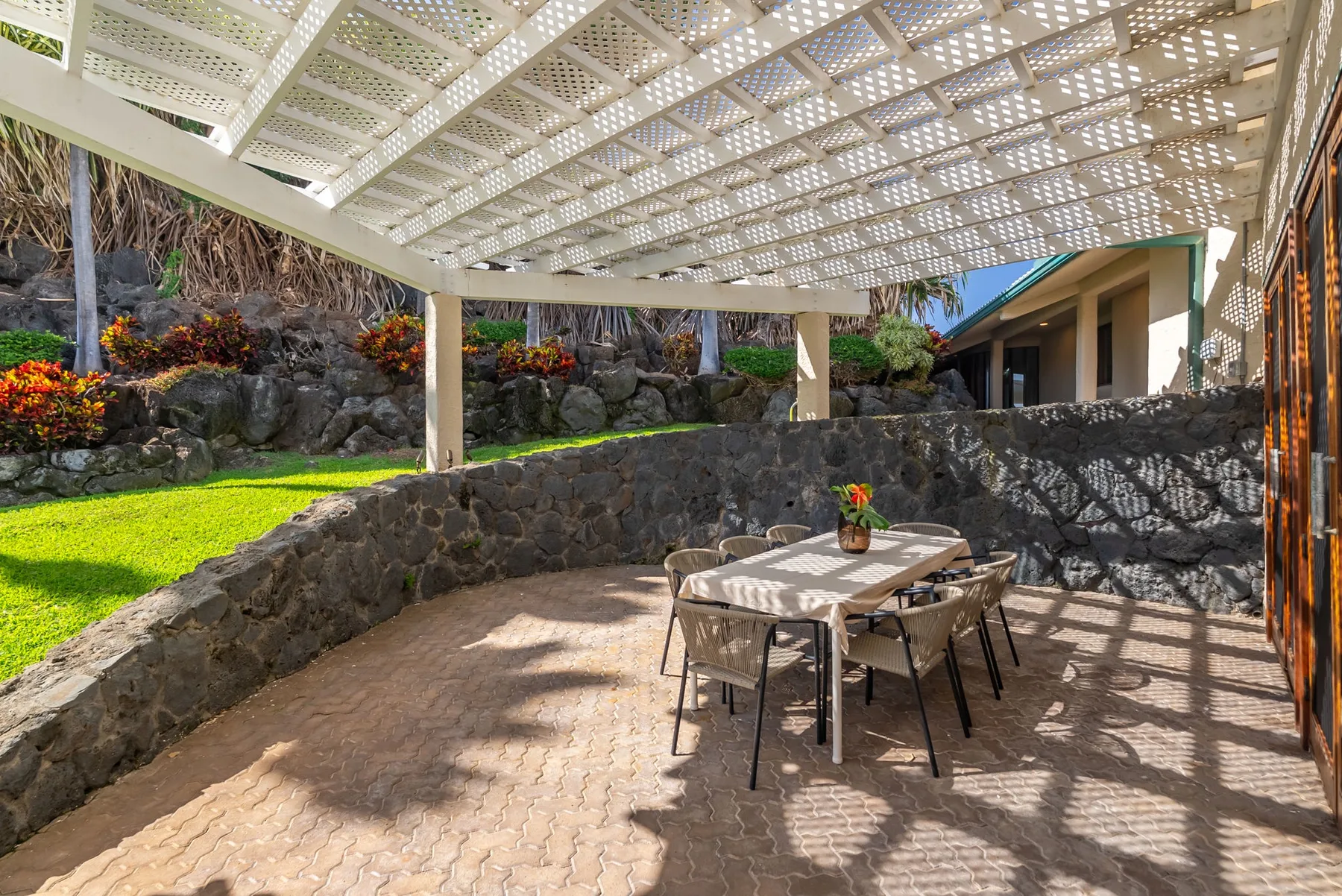 An outdoor dining patio sits beneath a white lattice pergola beside a stone wall.