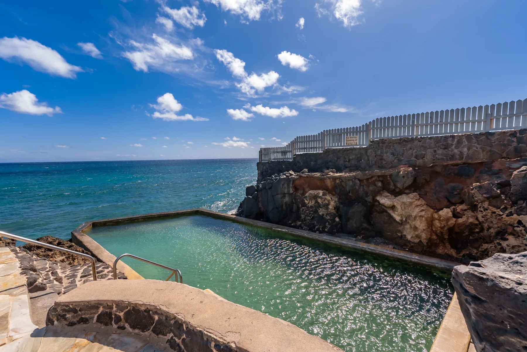 A seawater pool sits beside the rocky coast beneath a bright blue sky.