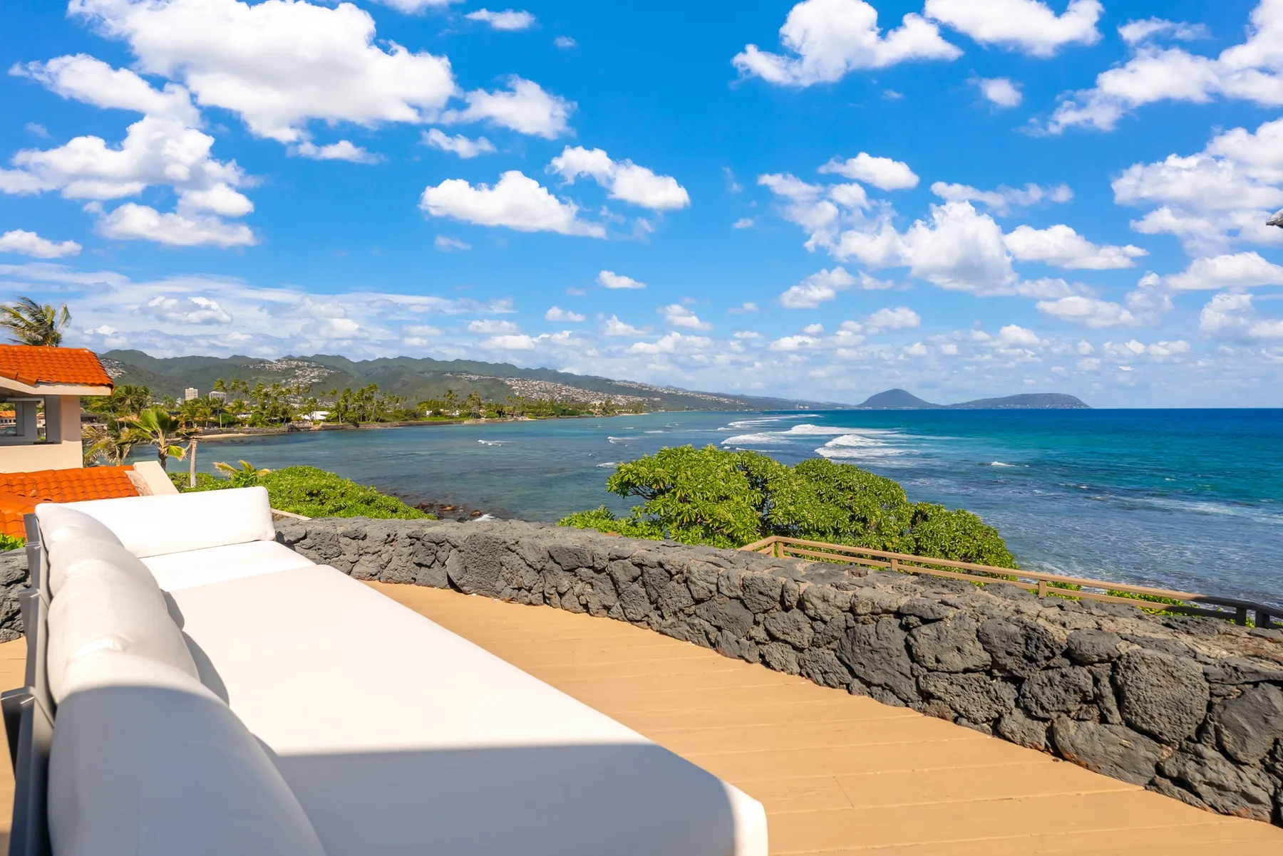 A seaside terrace overlooks the ocean, with cushioned seating and a low stone wall.