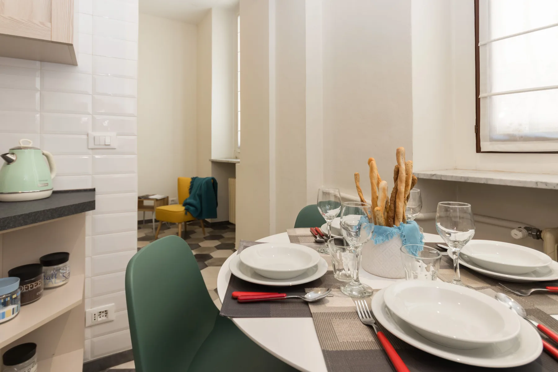 Una mesa de comedor está puesta con platos y copas junto a una cocina alicatada.