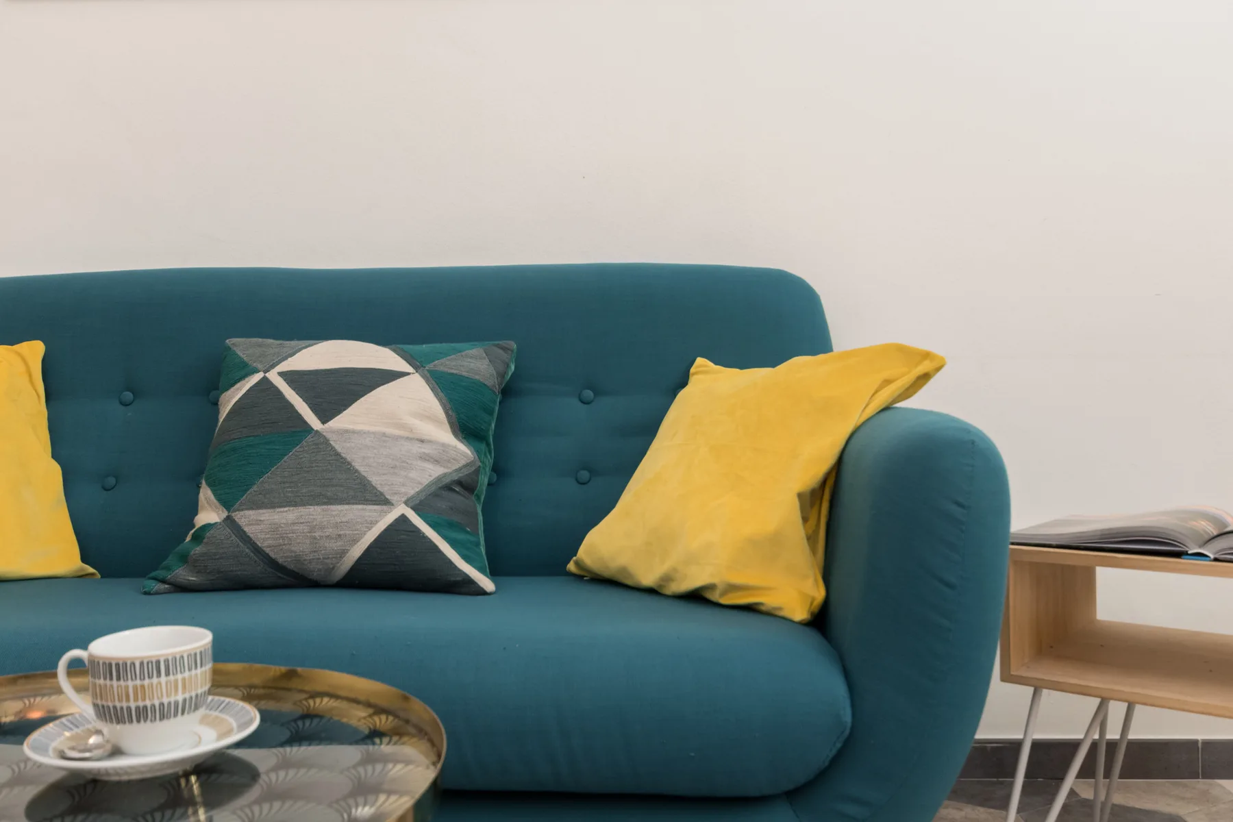 Un sofá verde azulado con cojines amarillos y estampados está junto a una mesa de centro de vidrio.
