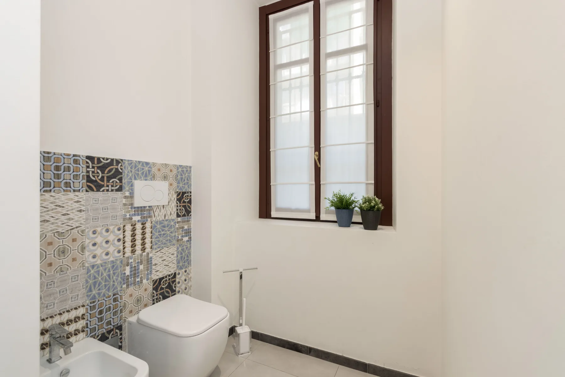Un baño luminoso con una pared de azulejos estampados y una ventana alta.